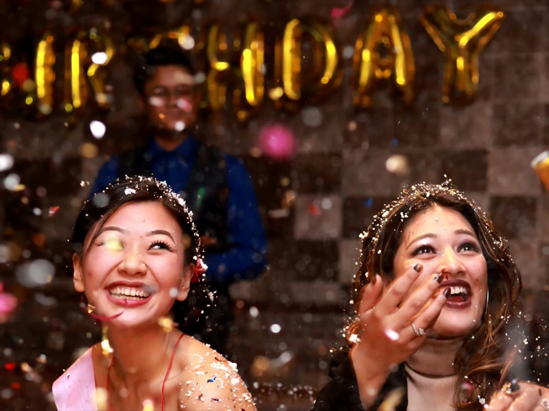 Deux amies éclatent de rire sous une pluie de confettis, devant des ballons dorés d'anniversaire