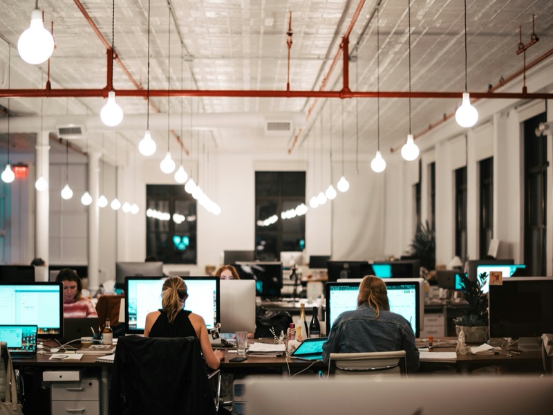 Un open space le soir, sous des ampoules suspendues : des collaboratrices travaillent à leur bureau